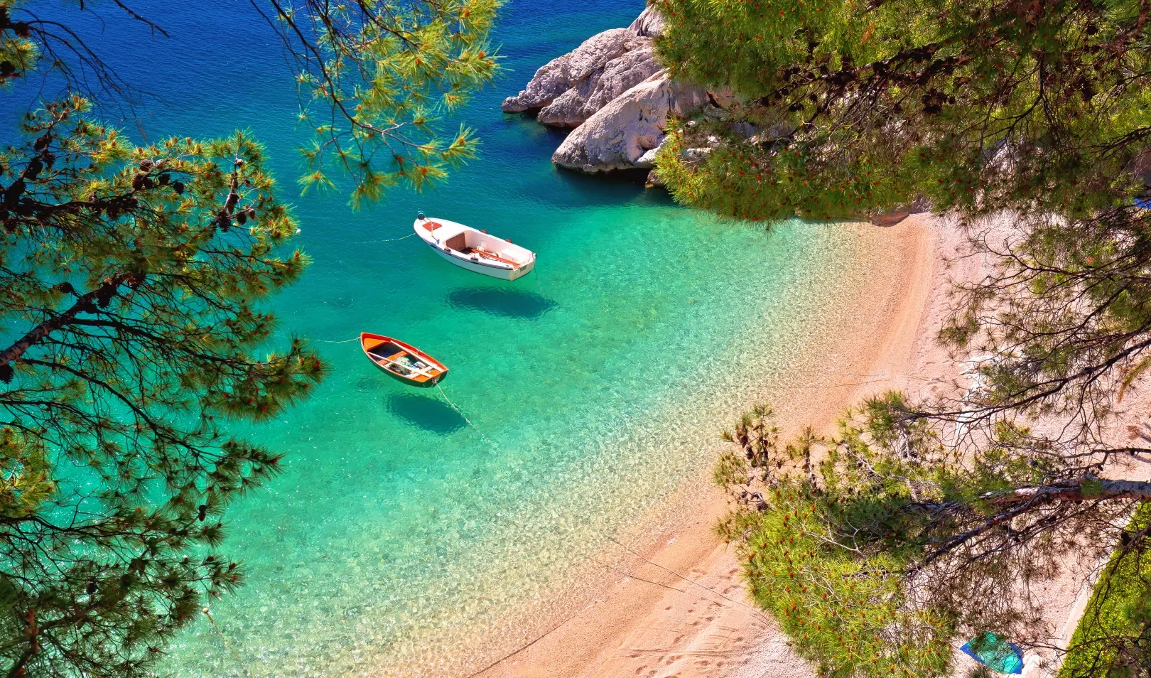 Pogled iz zraka na osamljenu šljunčanu plažu s tirkiznim morem, dva čamca na vodi i borovim granama.